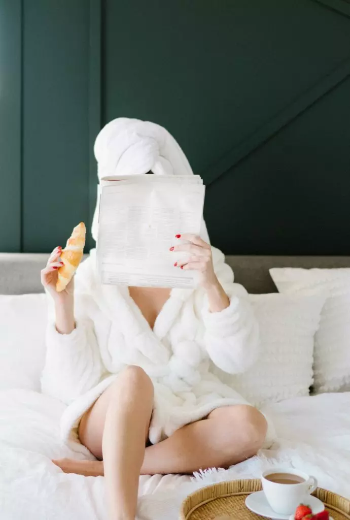 Woman in a bathrobe sitting in her bed, eating a croissant and leisurly reading a letter from the Mind Craft Archives