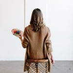 A woman with long hair in a brown knitted jacket holds a painter’s palette while studying a painting—illustrating Buying Art as both creativity and Smart Investment.