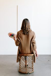 A woman with long hair in a brown knitted jacket holds a painter’s palette while studying a painting—illustrating Buying Art can be both passion and Smart Investment.