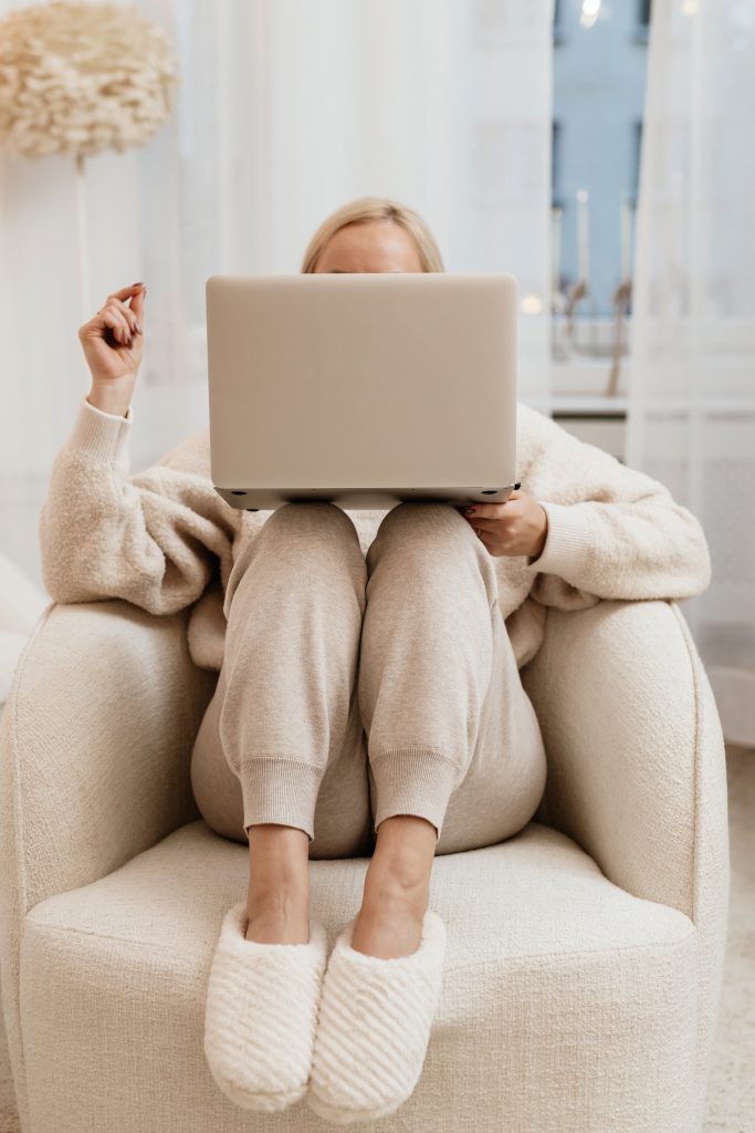 A woman sitting on a chair, dressed wearing a pair of cosy greige jogging trousers. Reading the Concoierge´s Selection on her Laptop.